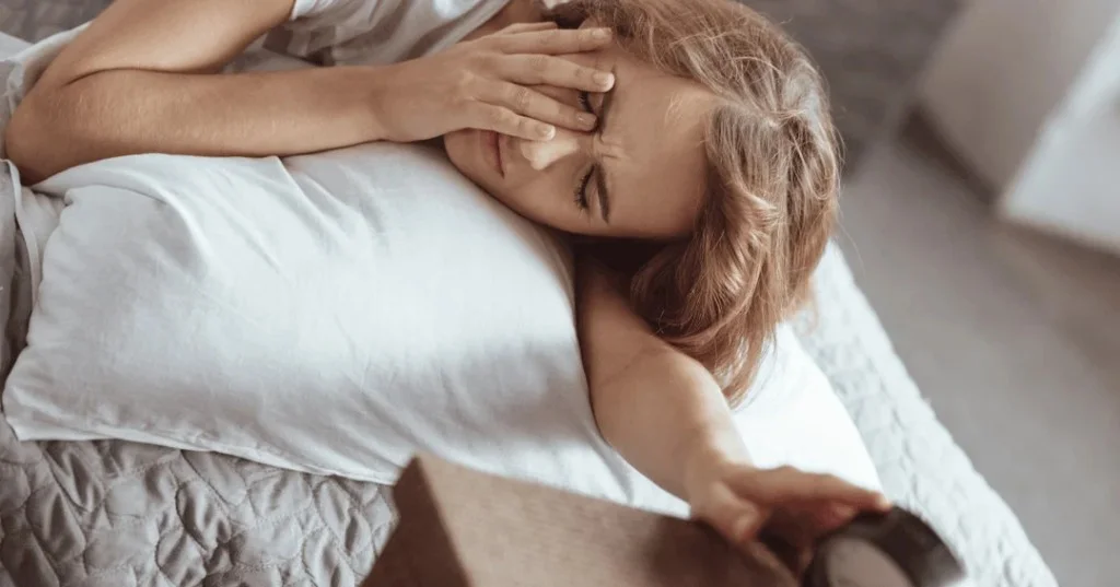 Woman groggy in bed reaching to turn off an alarm clock